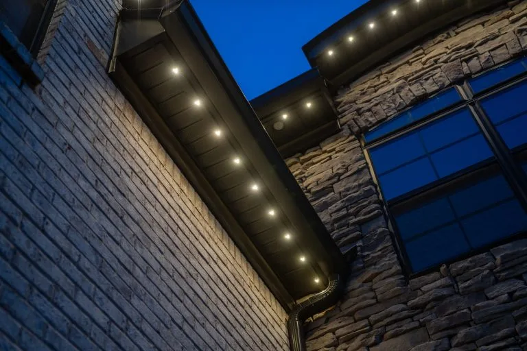 Warm lights illuminate the edge of a modern stone and brick house at dusk