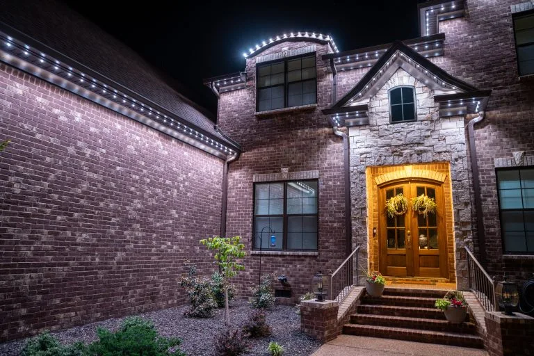 A warmly lit brick house features decorative lights and a welcoming front door