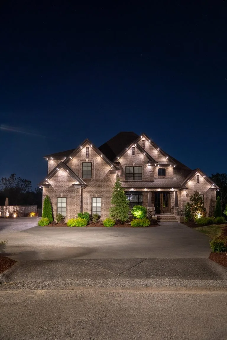 A beautifully lit house with festive lights, surrounded by lush greenery at night