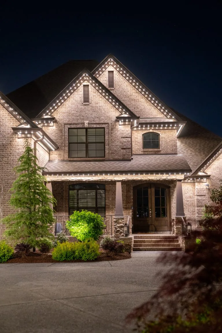 A beautifully lit brick house stands out against the night sky, surrounded by greenery