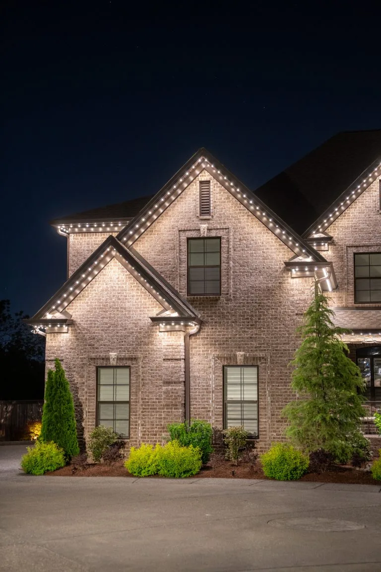 A beautifully illuminated brick house features landscaped greenery at night