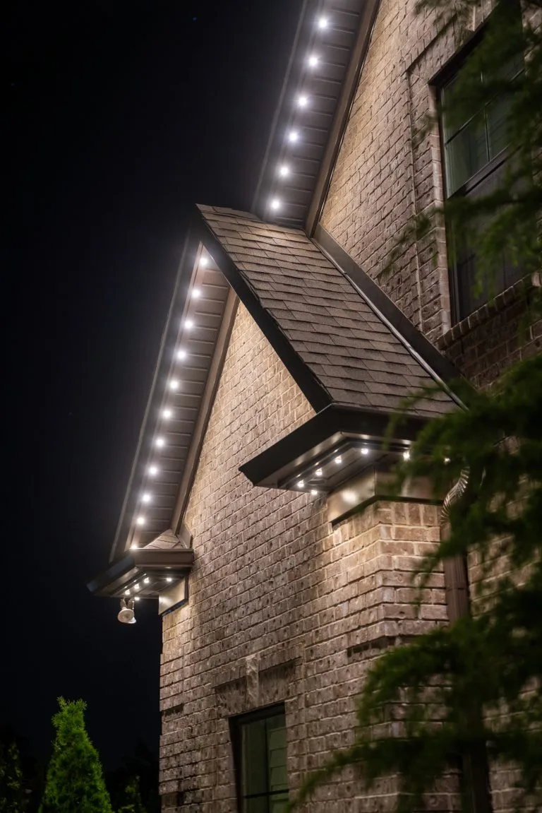 Warm, white lights outline the eaves of a brick house at night