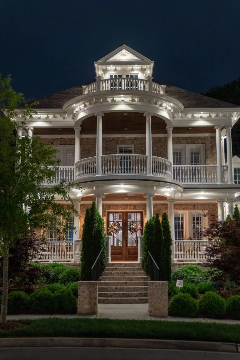 A grand, illuminated house features a spacious porch and lush greenery
