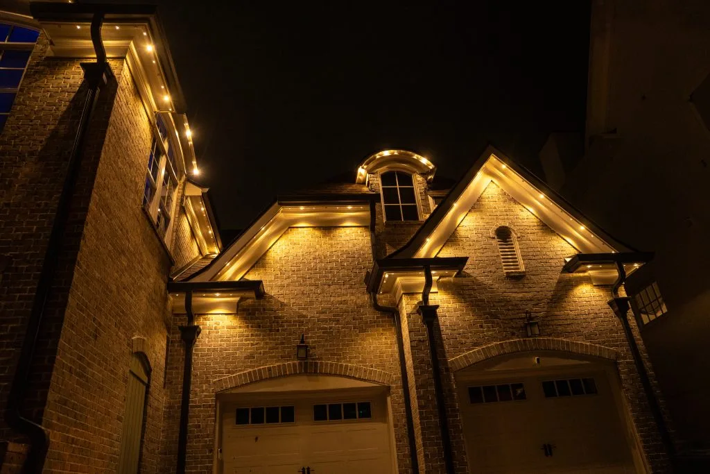 Warm, glowing lights outline a charming brick house under a dark night sky