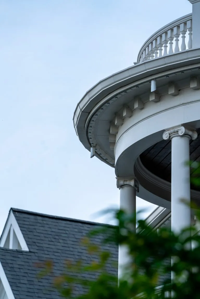 A round balcony with white columns overlooks a sloped roof under a blue sky