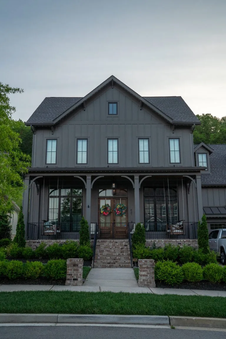 A modern, dark gray house features a welcoming porch and landscaped front yard