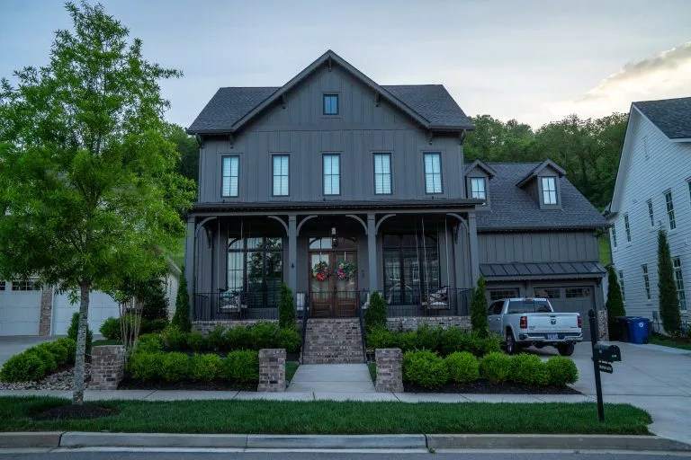 A large, modern house with a porch and lush greenery surrounds it