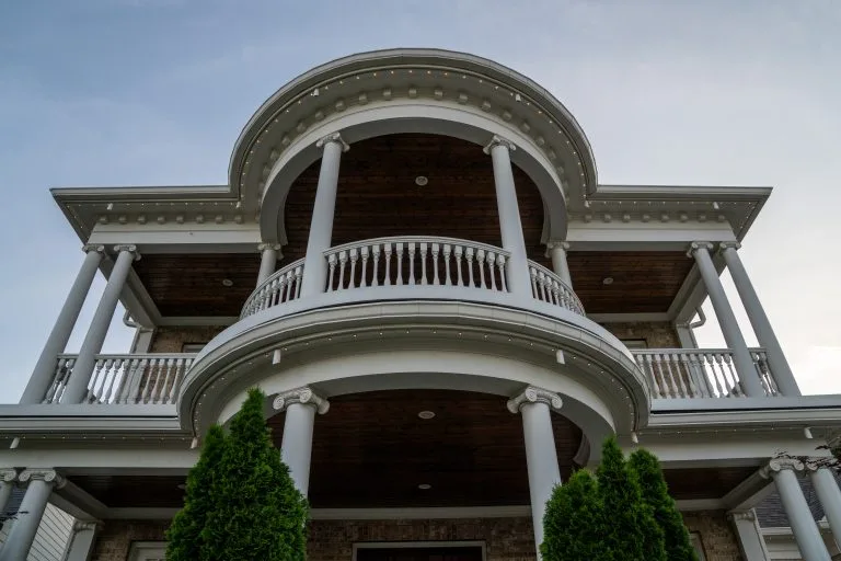 A grand house with a curved balcony and ornate columns rises against a clear sky