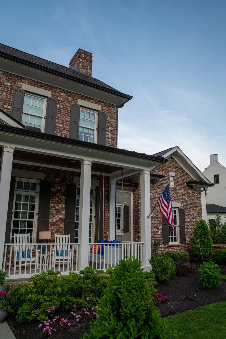 A charming brick house features a welcoming porch and flowing greenery, with an American flag displayed