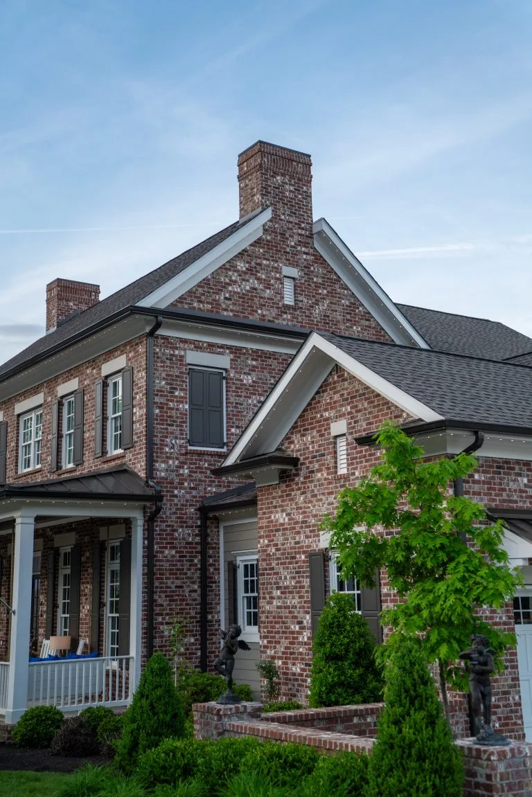 A charming brick house features a sloped roof and lush greenery surrounding it
