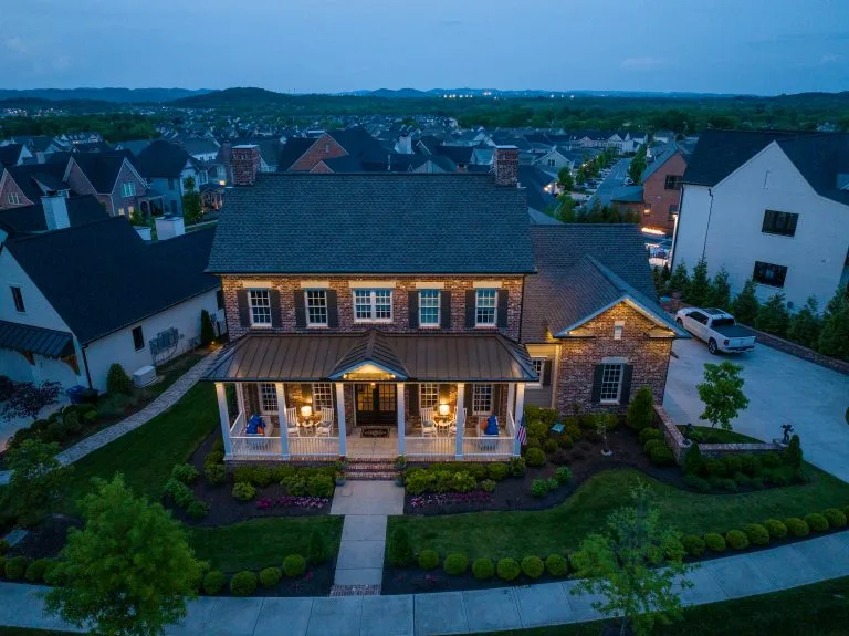 A charming two-story house with warm lights and a landscaped yard sits in a suburban neighborhood