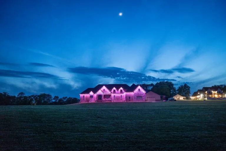 A brightly lit house shines under a twilight sky with scattered clouds