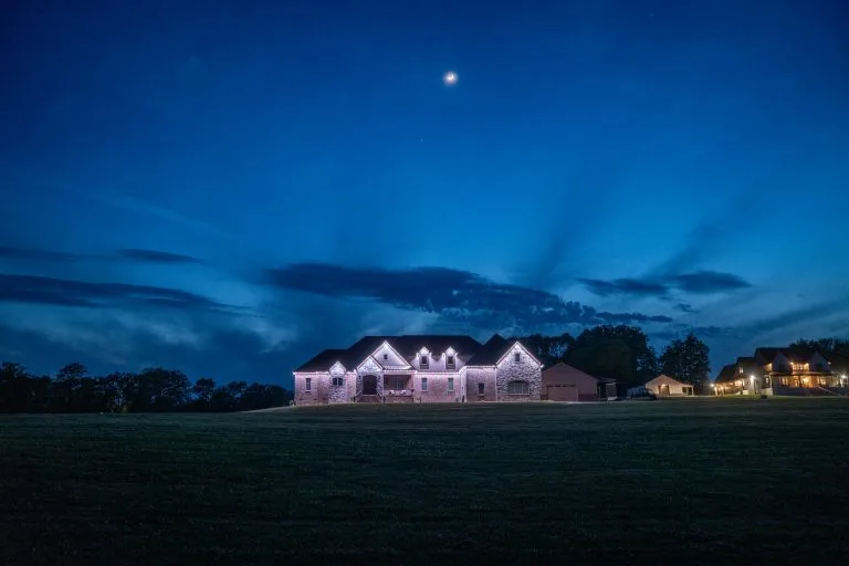 A large, illuminated house stands against a twilight sky with scattered clouds and a visible moon
