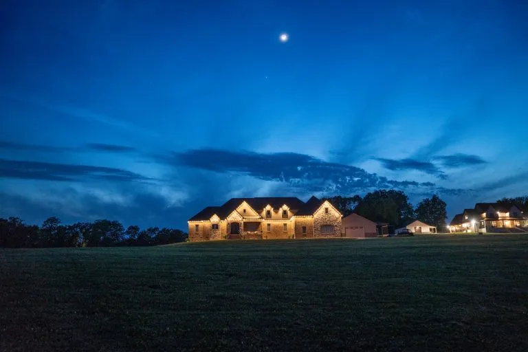 A beautifully lit house stands against a twilight sky with scattered clouds and a crescent moon