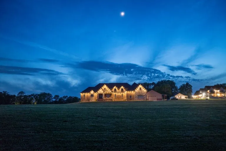 A beautifully lit house stands under a twilight sky with scattered clouds and a crescent moon