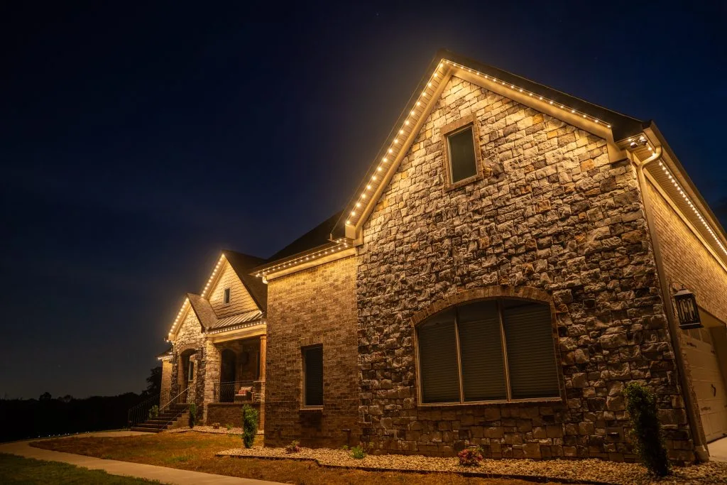 Roofline lighting enhances the warm glow of a stone house against a dark night sky