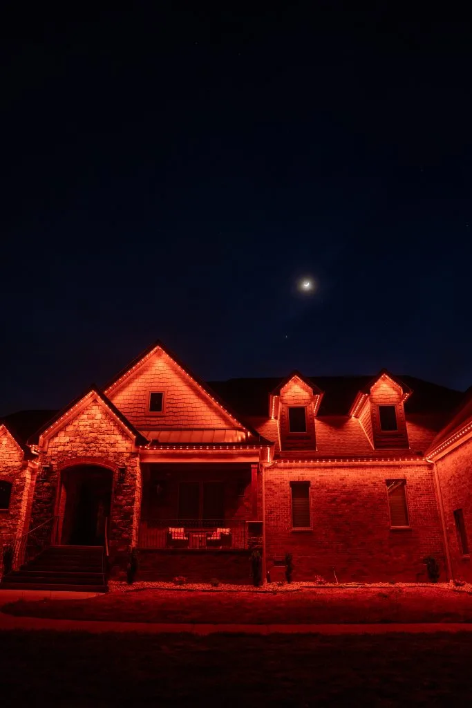 A beautifully lit brick house glows warmly against a dark night sky with a crescent moon