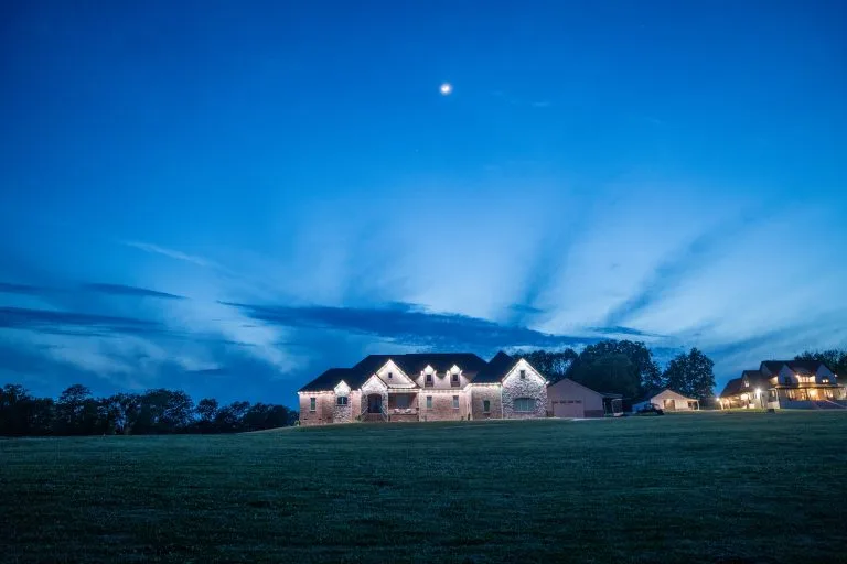 A large house glows warmly under a crescent moon and vibrant twilight sky