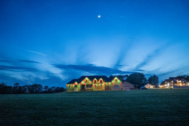 A large, illuminated house stands against a twilight sky with wispy clouds and a crescent moon