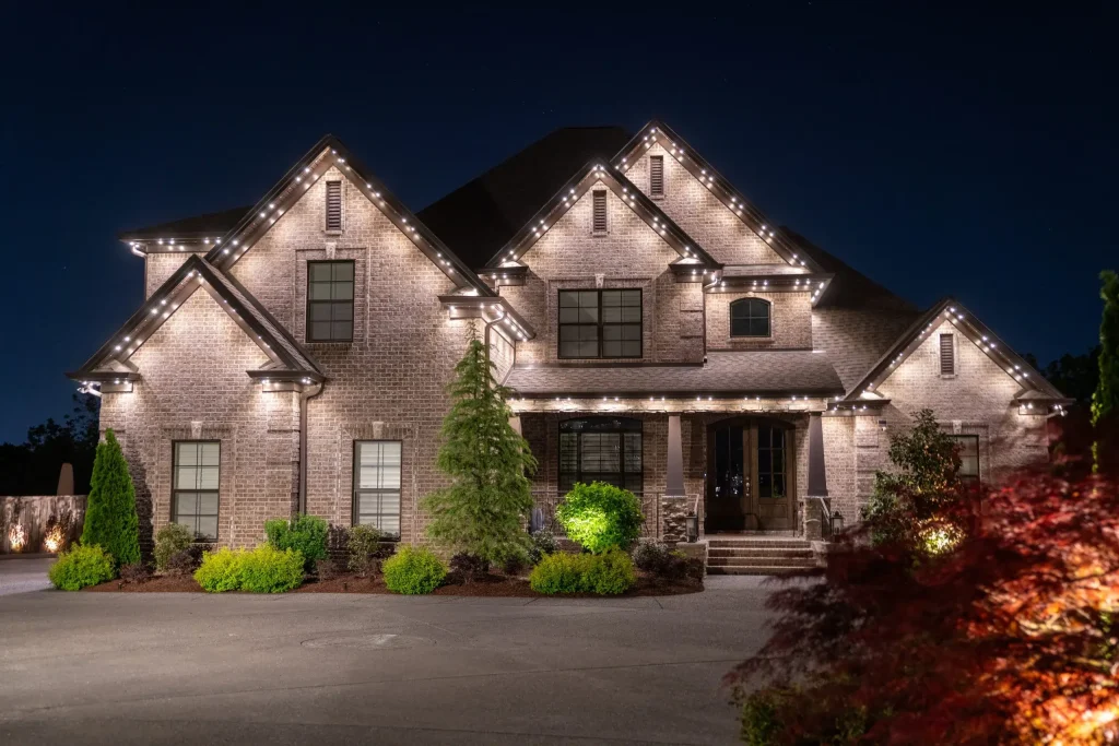 A beautifully lit brick house stands elegantly at night, surrounded by lush greenery